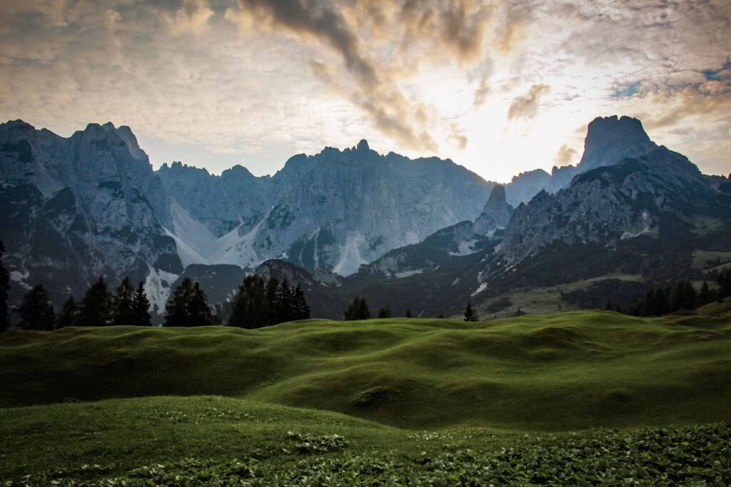 Majestätische Alpenlandschaft bei Sonnenuntergang, grüne Hügel und felsige Gipfel im Hintergrund.