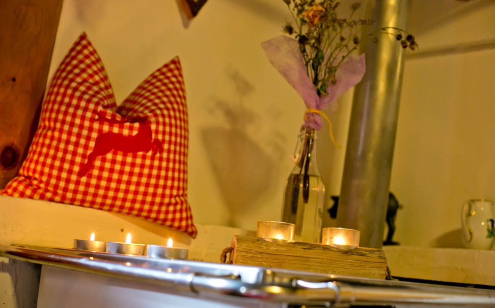 Gemütliche Alm-Einrichtung mit kariertem Kissen, Kerzen und blühendem Grün.